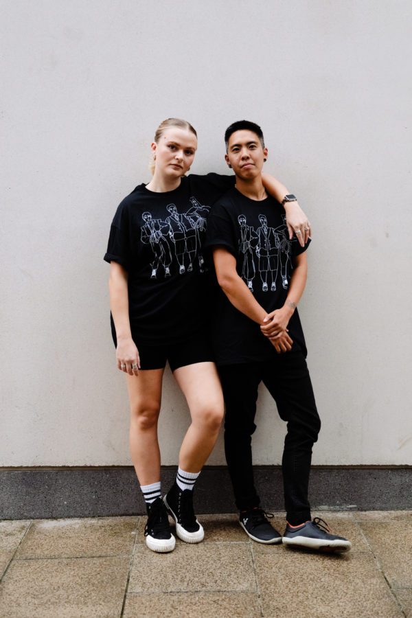 two people wear short sleeved black t shirts with white line illustration on them of 3 queer people