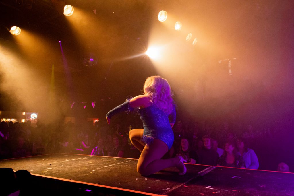 Drag queen Ivy Alexander kneels on a catwalk under bright golden lights. Ivy is a plus sized queen with big blonde wig and wears a blue sequin bodysuit