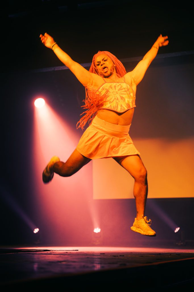 Angel Blaque leaps onstage under pink lighting. Angel is a Black, femme with pink braids wearing a pale crop top and cute flared mini skirt with chunky white trainers.