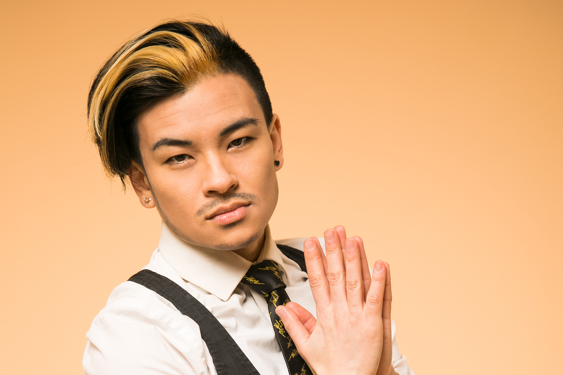 a youthful East Asian drag king stands with his hands together in front of his chest. He is captured sideways onto the camera. He wears a white shirt with balck waistcoat and black jeans with black sequin detailing. He has a thin moustache and black hair with bleach blonde streak worn in a long side quiff.
