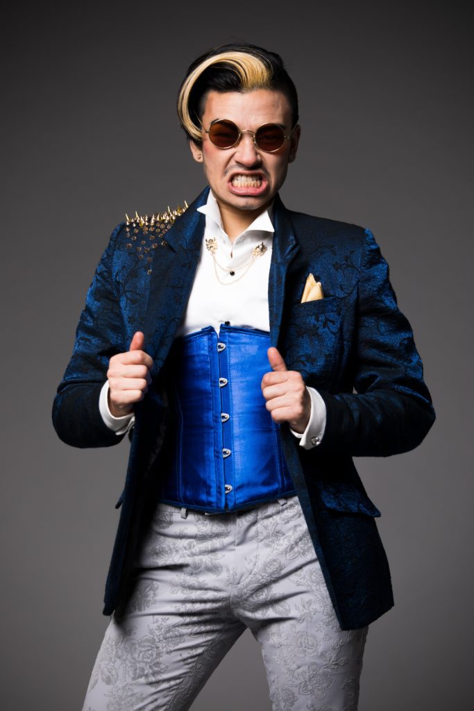 Drag King Sigi Moonlight, a slim East Asian masc guy is pictured bearing his teeth in a fierce grimmace. He wears a white shirt with royal blue satin waist corset and dark blue brocade jacket with gold embellishments including spiked studs on the right shoulder. He also wears grey brocade trousers and dark round sunglasses. His hair is black with a white streak and styled into a side quiff.