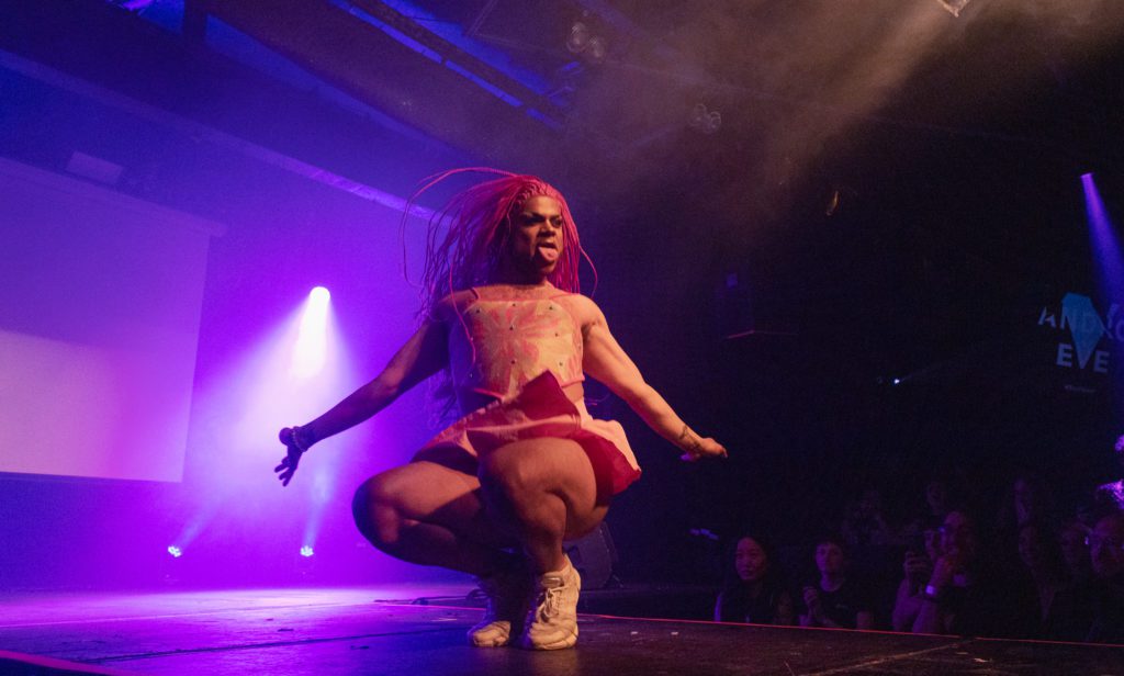 Angel Blaque duck walks onstage under pink lighting. Angel is a Black, femme with pink braids wearing a pale crop top and cute flared mini skirt with chunky white trainers.