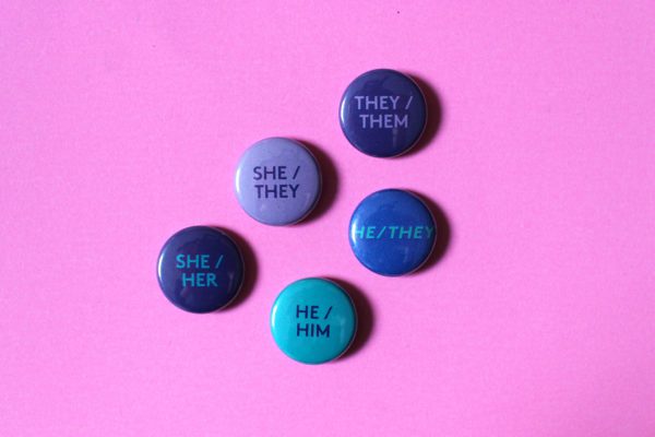 5 badges of different designs sit on a pink surface. One is lilac with the words ‘She / They’ in purple printed on, another is purple with ‘They / Them’ printed on in lilac. Another is turquoise with ‘He / HIm’ printed in dark blue, another is dark blue with ‘He / They’ in turquoise and another is purple with ‘She / Her’ in turquoise.
