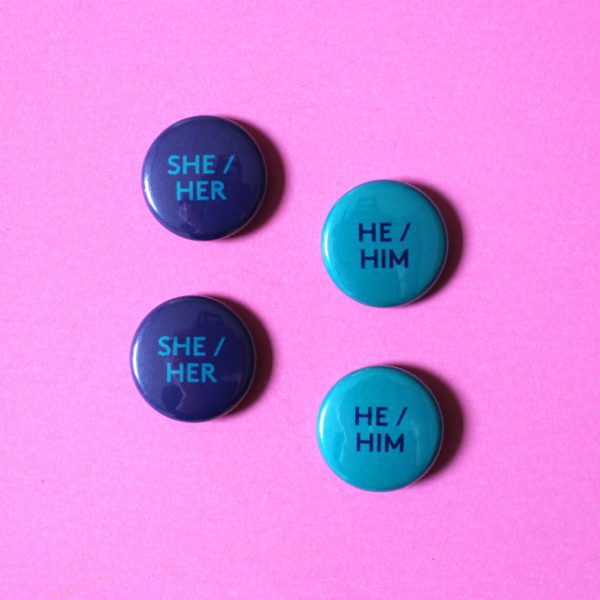 4 circular badges sit on a pink surface. There are two designs that are purple with the words ‘She / Her printed in turquoise, and two designs that are turquoise with ‘He / HIm’ printed in dark blue.