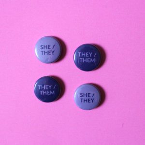 4 circular badges sit on a pink surface. There are two designs that are purple with the words ‘They / Them printed in lilac, and two designs that are lilac with the words She / They in purple.
