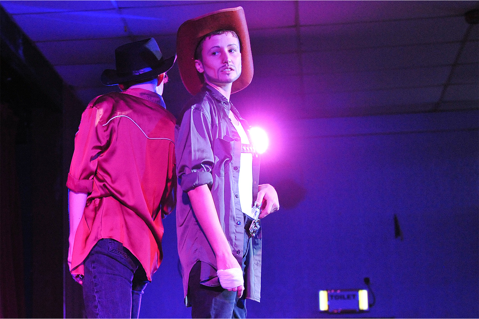 Two slim, white drag kings dressed like cowboys with cowboy hats and shirts, stand back to back on a stage. They are lip syncing and lit by blue, red and purple lighting.