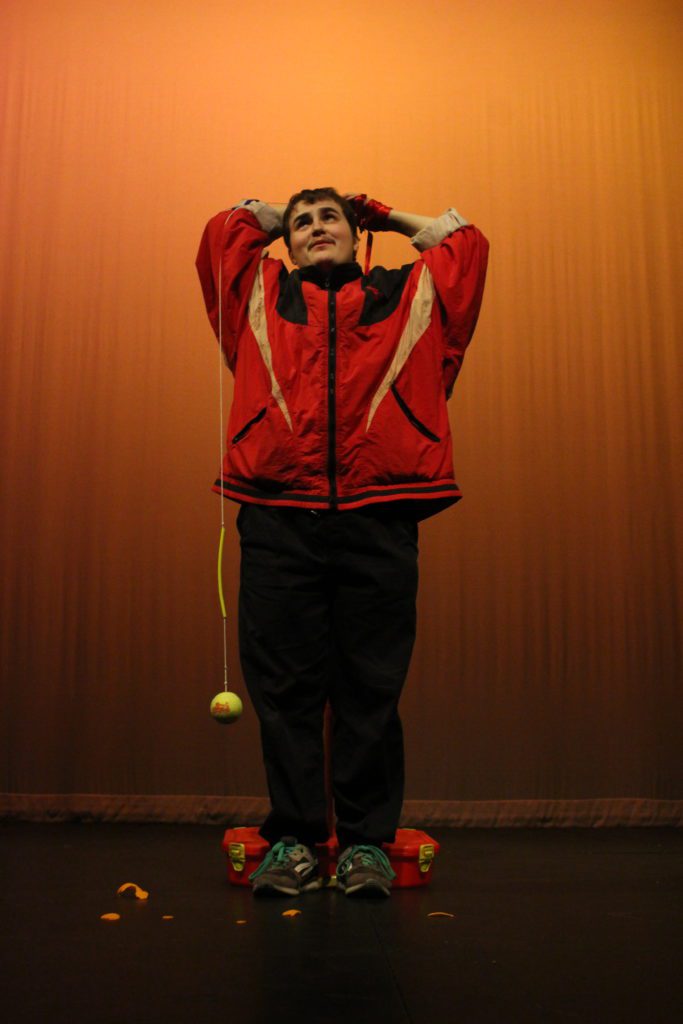 a white woman with short. dark hair wearing a red sporting zip up top holds her arms above her head. Attached to her waist is a piece of string with a yellow tennis ball attached