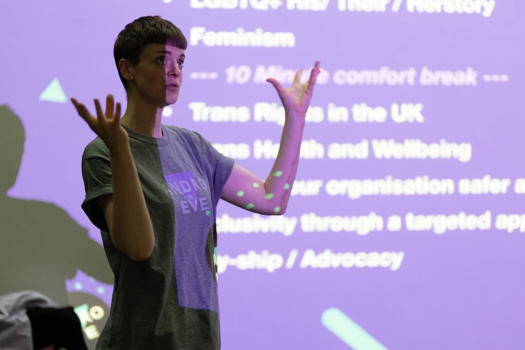 Finn, a slim white androgynous person with short hair is speaking in front of a screen with a session plan projected onto it