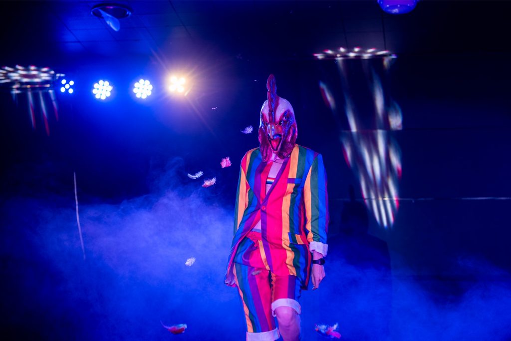 Toby Carvery wearing a rainbow suit and chicken head, struts on stage