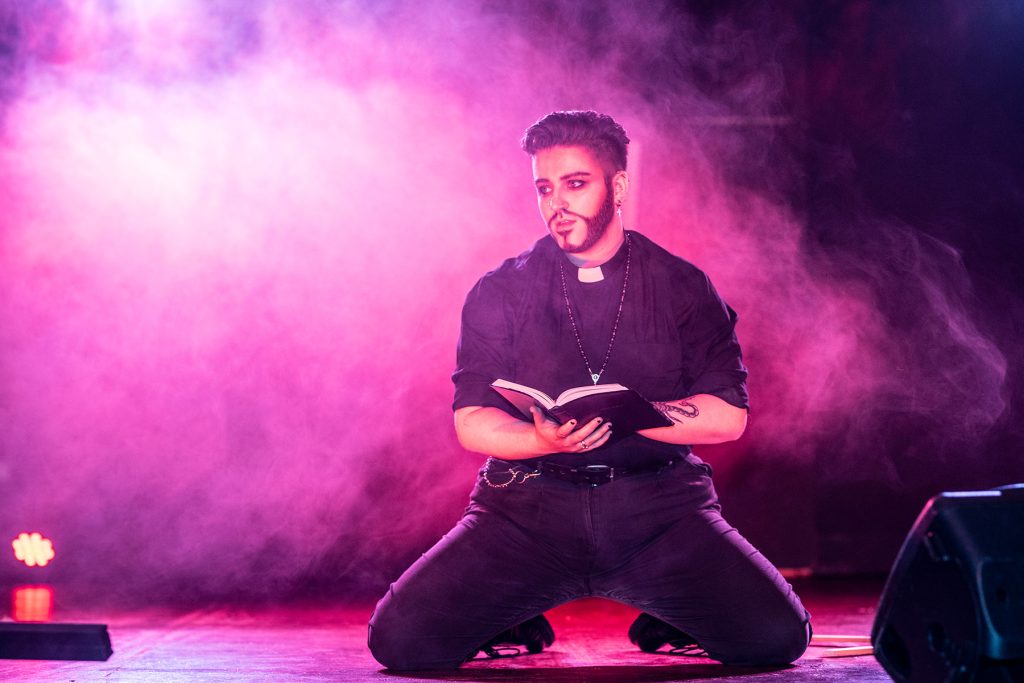 Billy Butch, a white drag king with swept back dark hair, is dressed as a priest in black with a dog collar. He is kneeling on stage holding the bible