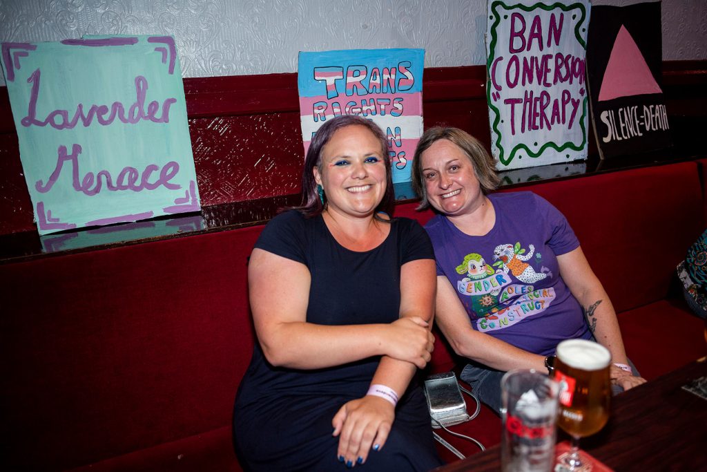 Two white people sit next to each other on a red velvet banquet seating smiling. Behind them are handmade card placards that say ' Lavender Menace' 'ban conversion therapy'