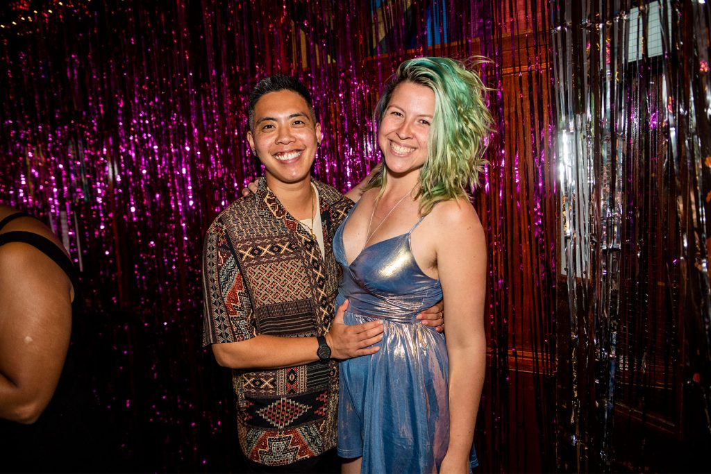 A Chinese man and white femme with blue hair stand with their arms around each other smiling in front of a pink glitter curtain