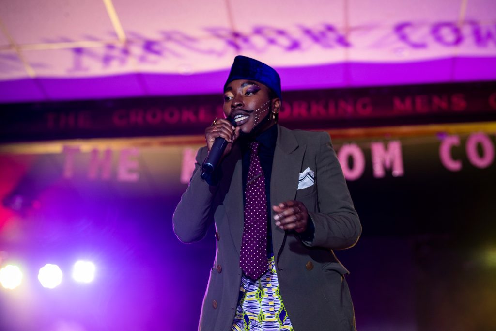 Wesley Dykes, a Black West African drag king stands singing into a mic on stage lit by purple lighting.