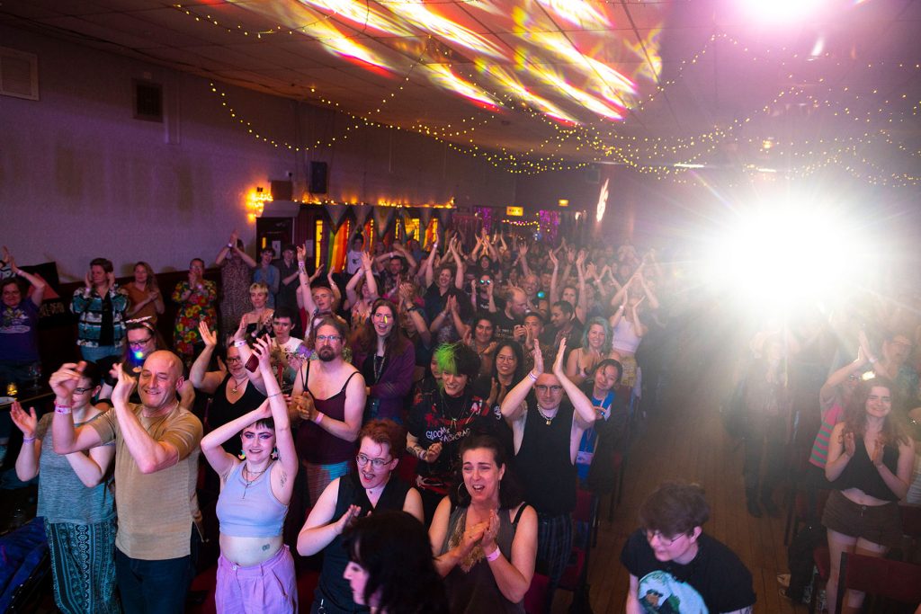 A crowd of people cheers and claps in a big hall lit by warm lighting.
