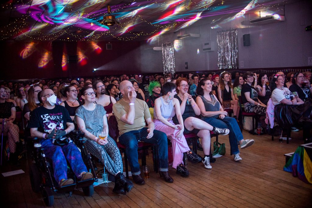 A crowd of people sit looking upwards at a stage in a hall lit by rainbow coloured lights