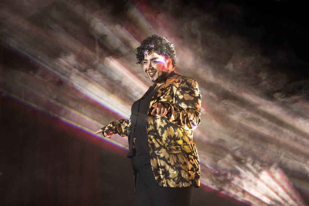 Shardeazy Afrodesiak, a mixed race drag ting with strong beard and quiff smiles onstage wearing a gold and black blazer