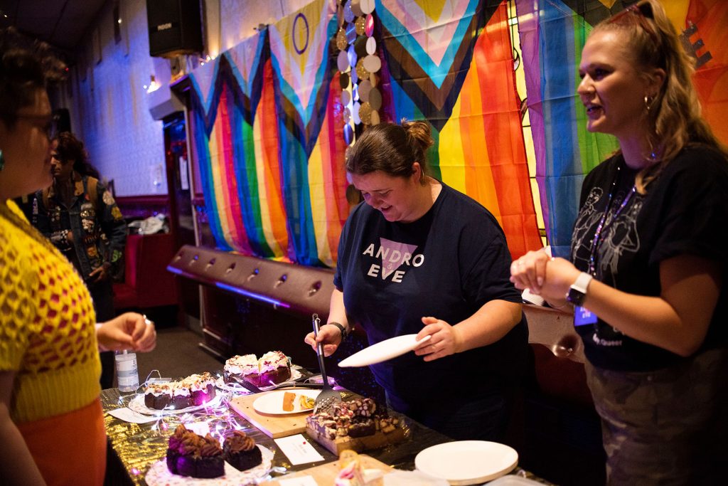 two people stand behind a cake stall serving cake with Pride flags behind them