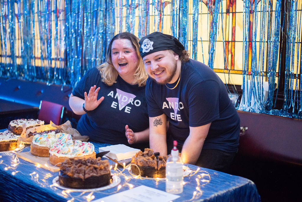 Two white queer people smile wearing Andro and Eve Tees as they sell tasty cake