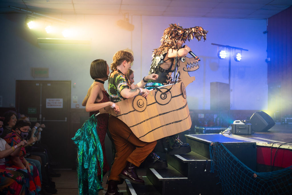 Drag King Oedipussi dressed as a viking, charges up the stage steps with 3 people in his cardboard boat