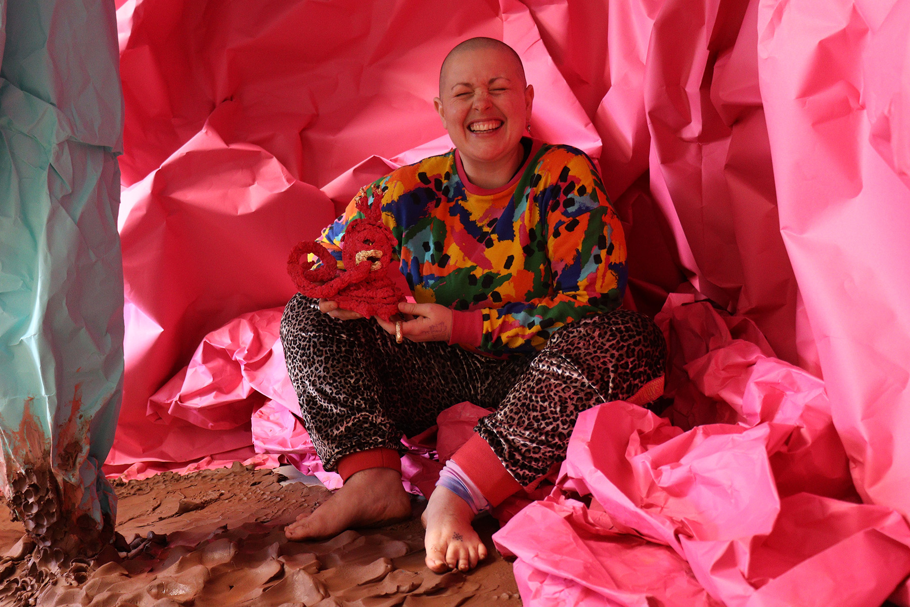 Colour photo, interior. Kitt, a white, shaven headed person with a wide crinkly nosed smile, sits amongst vast sheets of pink and mint green paper crumple to form a cave - like structure around them. on the floor is wet, raw terracotta clay in rolling folds.
