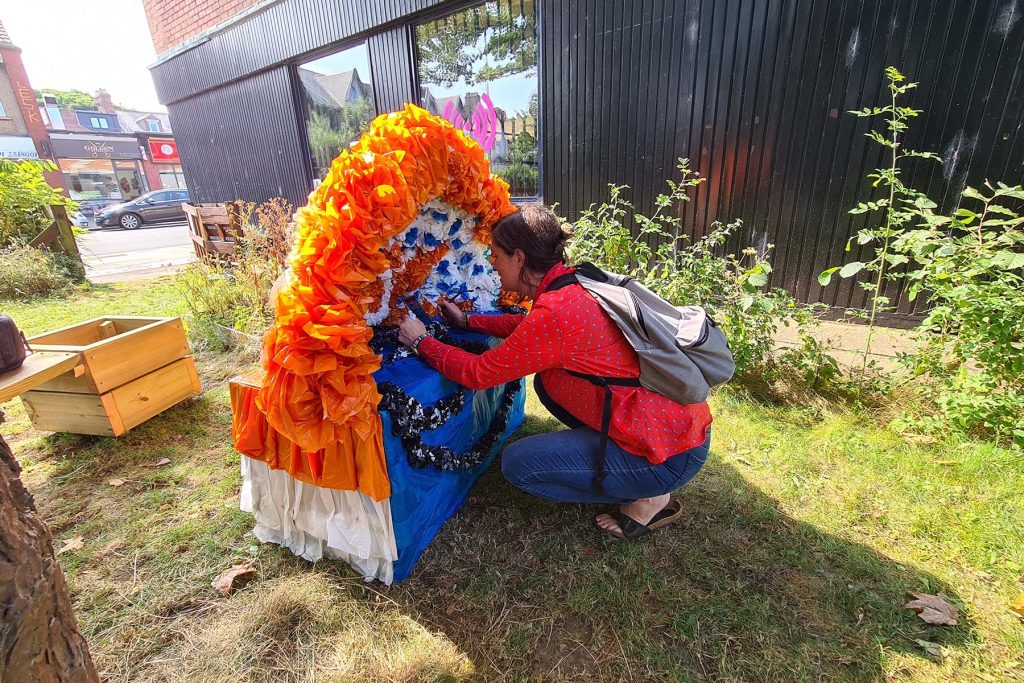 Colour photo, exterior. A shrine made from garlands of reused a soft plastics in blue, white, and orange sits on the floor in a patch of grass, one person is knelt down interacting with the shrine.
