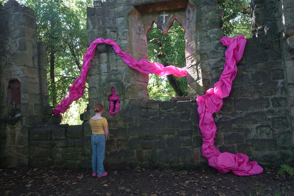 exterior. St marys chapel (a ruined chapel with part of an arch and several windows visible), woven through the ruins are bright pink, purple, blue and green lengths of crumpled, sculpted fabric. A young person stands and closely inspects the woven fabric. 