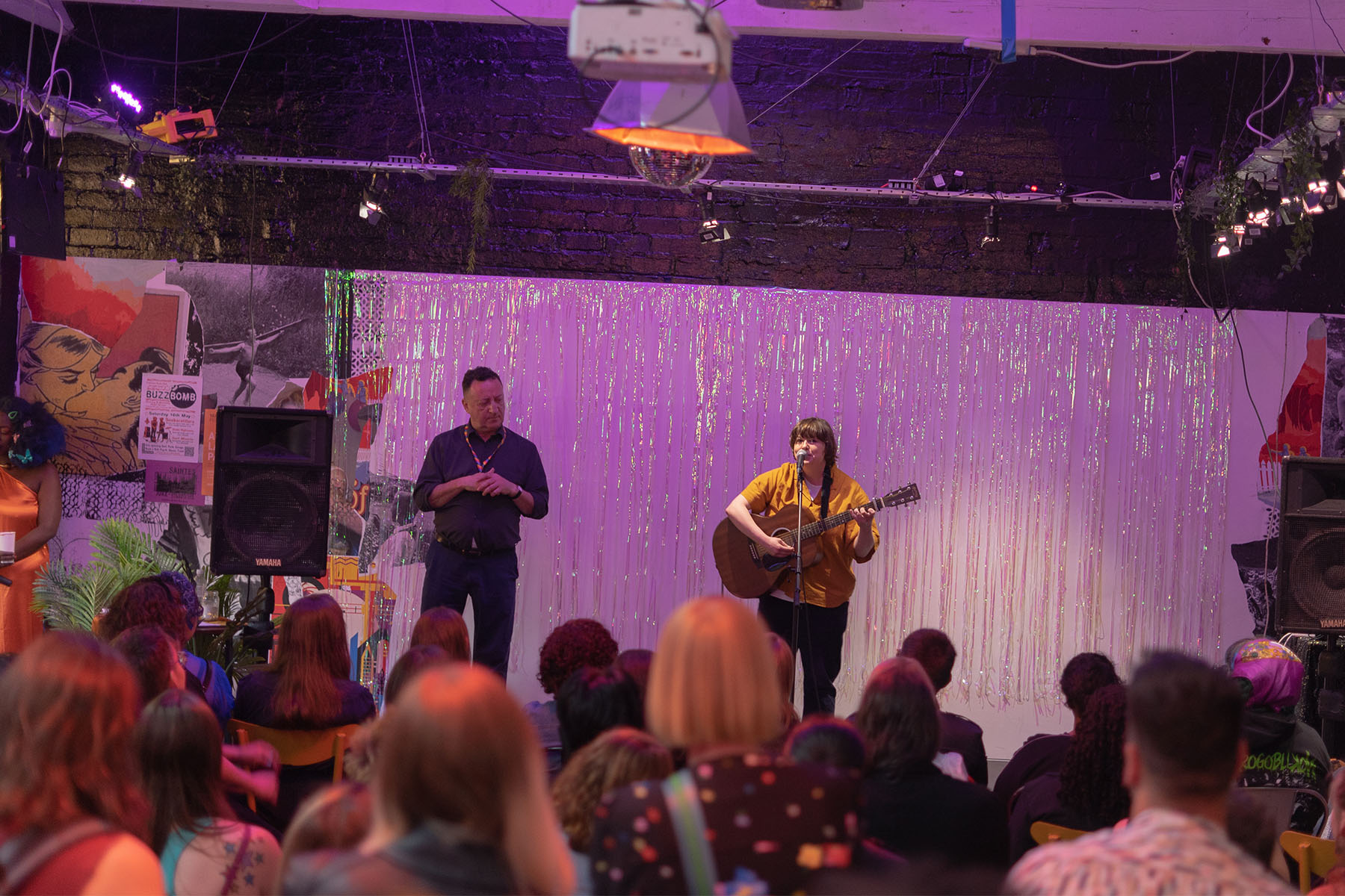 a white queer woman with short brown hair sings on a mic with a guitar in front of a seated audience