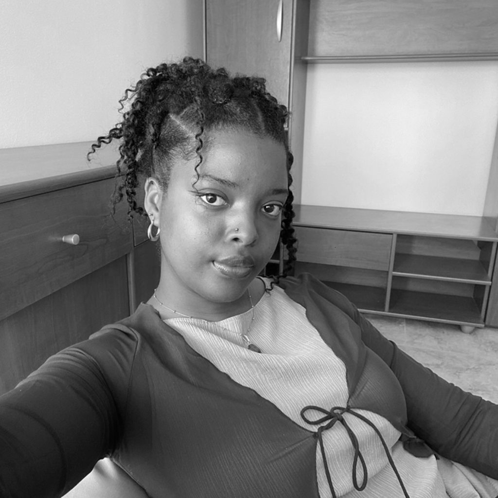 A black woman with twists sits in a room with wooden furniture, looking directly at the camera. She wears a beige dress and brown top and gold hoop earrings.