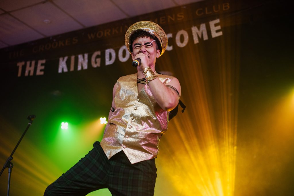 Drag king Jamie Fuxx in gold waistcast and matching cap and black trousers sings on the mic