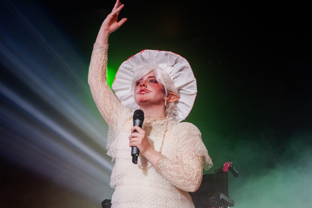 Drag king Pure Gliss sings dressed as a mushroom while onstage using a wheelchair