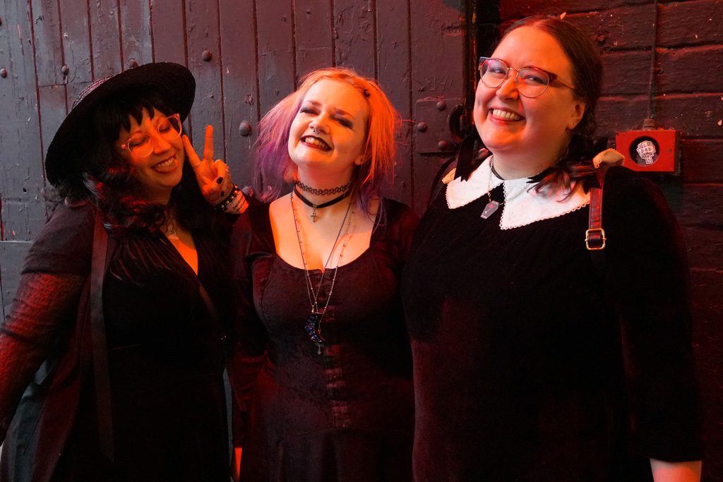 Three white people smile and pose wearing gothic outfits