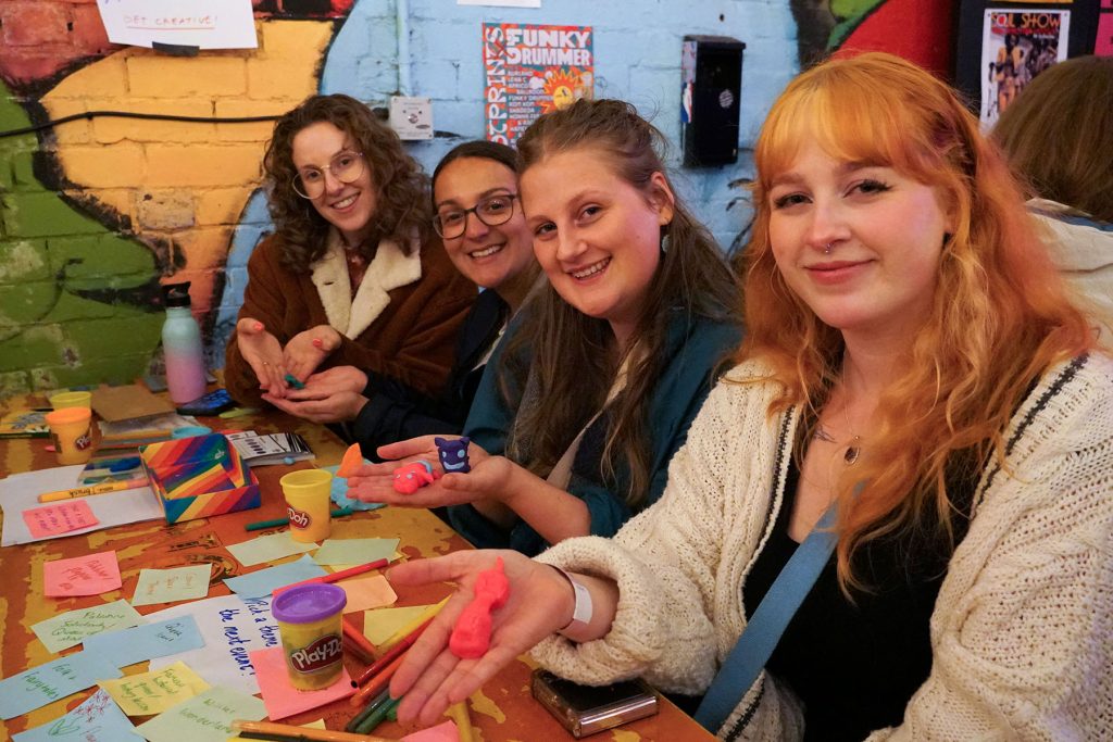 Four people hold their crafted sculptures seated at a table and smile