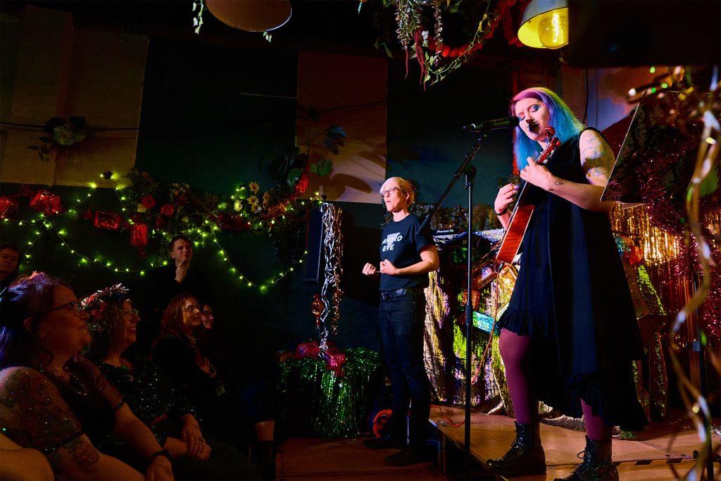 AMereKat, a white person with long blue hair plays a Ukelele on a small stage with Becky the BSL interpreter beside them