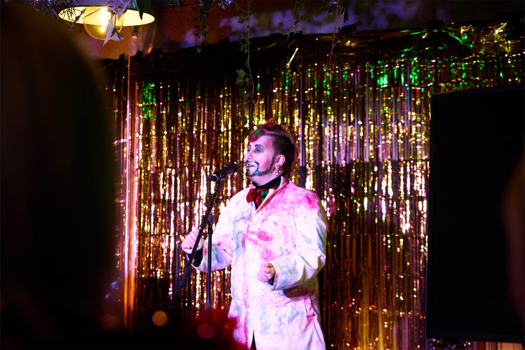 Drag king Raymond Petty, sings onstage wearing a lab coat. He is a white king with painted red beared.