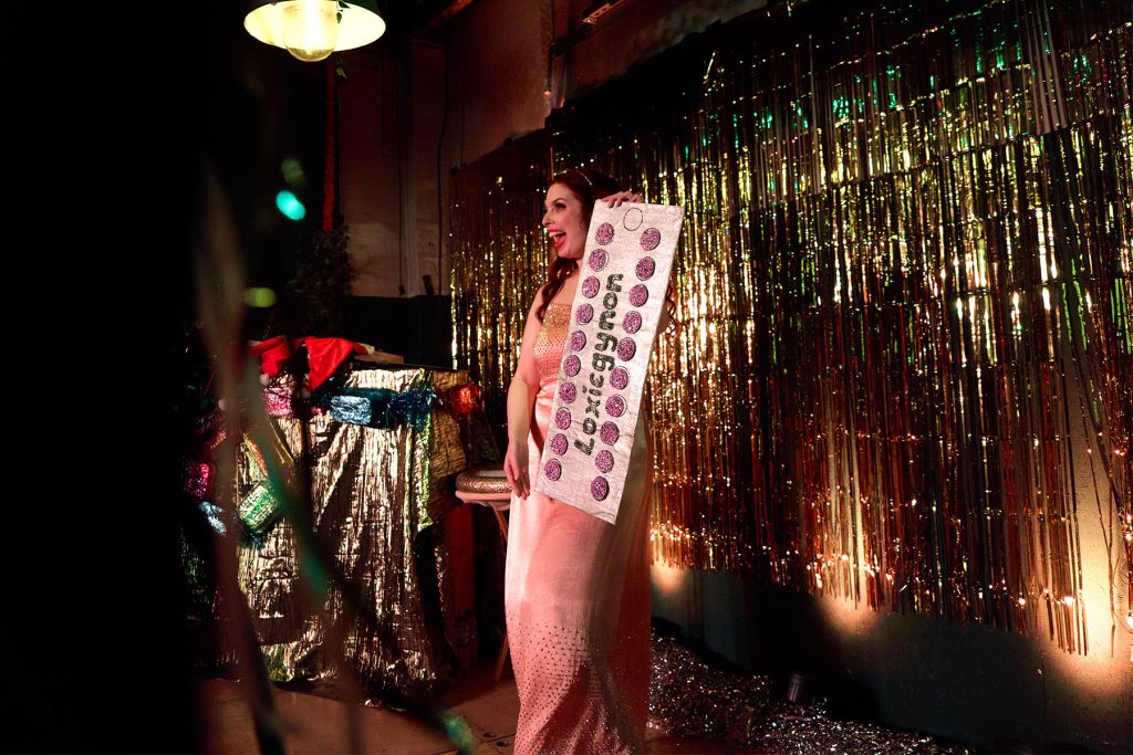 Loxie Tocin, a white glamorous redhead performer holds a giant contraceptive pill packet prop while smiling