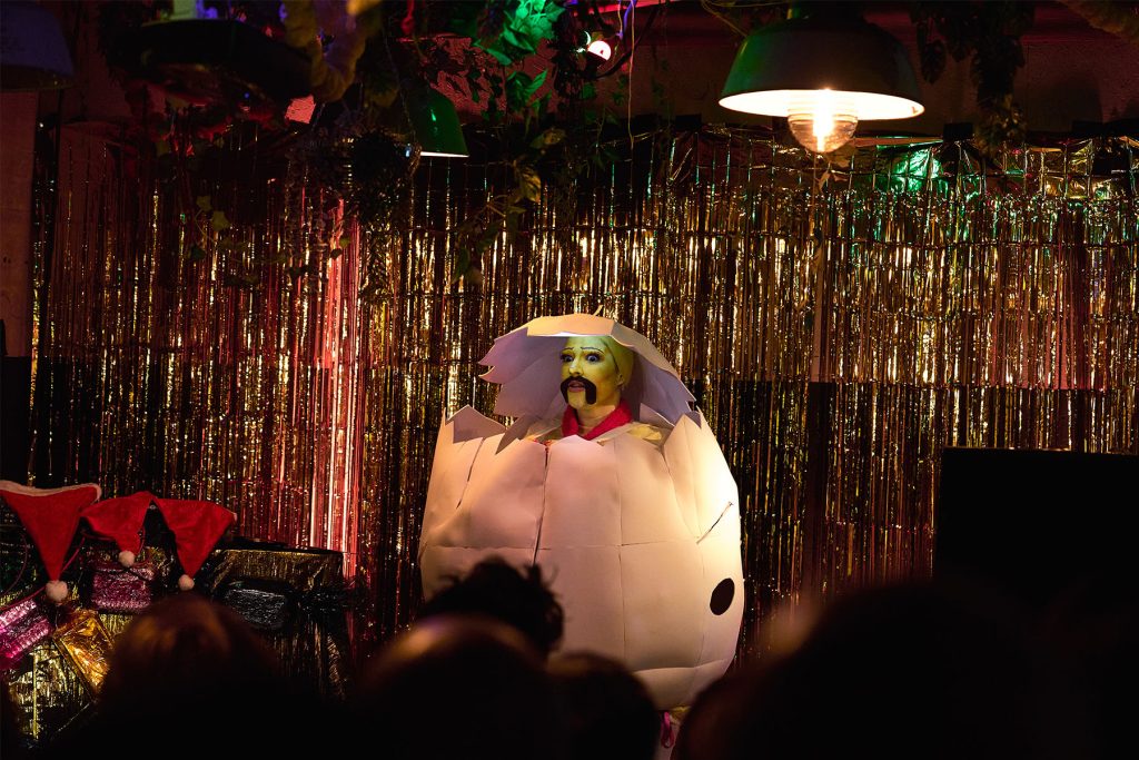 Hot Pot a white drag artist is onstage dressed as a chicken with a moustache inside a giant white egg