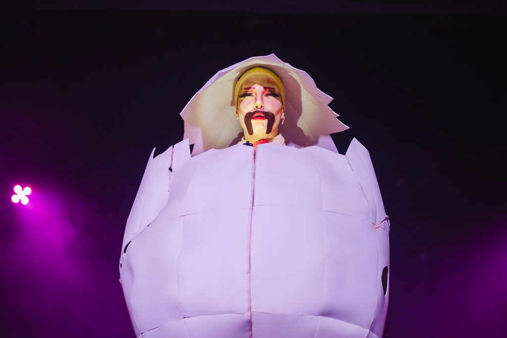White drag king Hot Pot lipsyncs with a yellow face inside a giant white egg