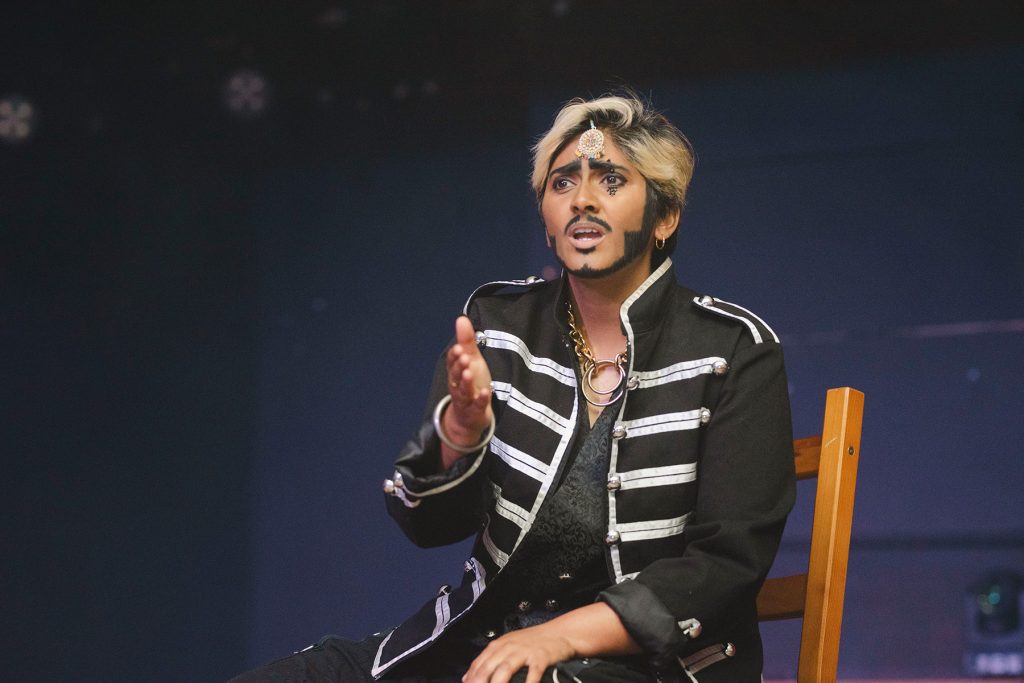 South Asian drag king Harddeep Singh lipsyncs seated onstage wearing military jacket