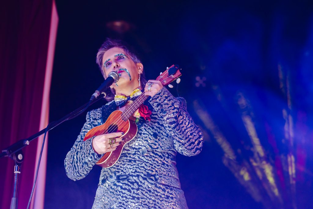 Blind, white drag king Tito Bone sings on a mic playing the ukelele.