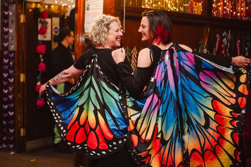 Two white women smile with their backs to camera showing off colourful butterfly wing capes