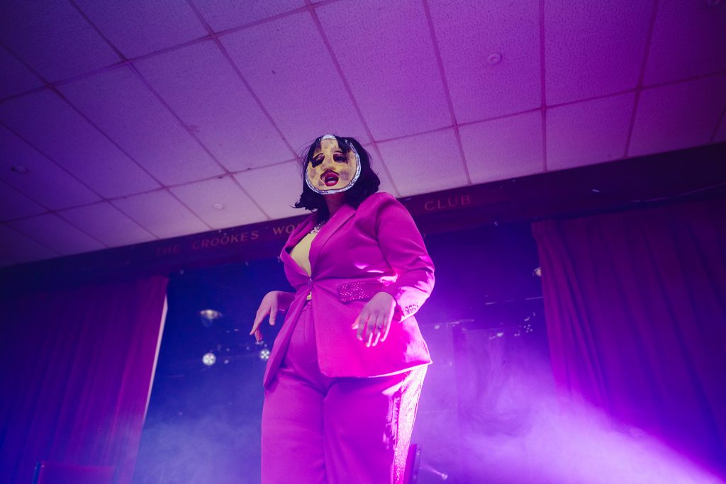 Hot Pot dressed in pink business suit with a quiche mask and brown wig