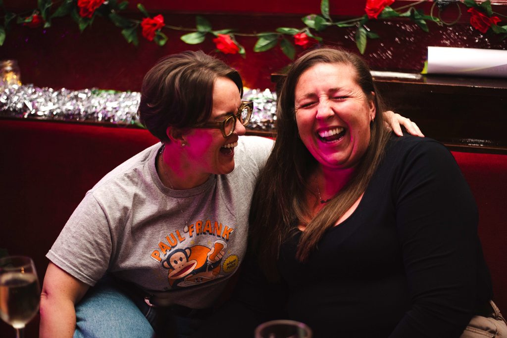 Two white people laugh with scrunched eyes and their arms around one another