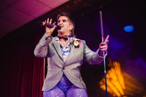 King Tito Bone sings on the mic holding his walking cane. Tito is w hite drag king with a grey mullet and turquoise glitter beard