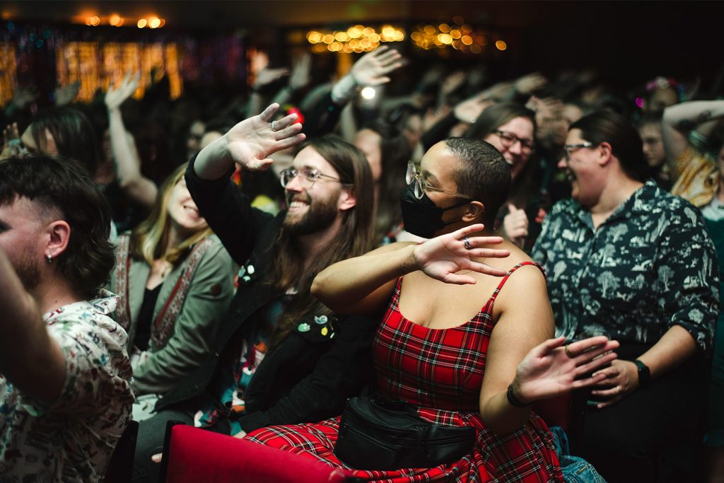 A diverse audiences smiles and waves their arms in the air