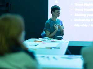 Finn, a slim androgynous person with short brown hair sits in front of a screen presenting to a group of people