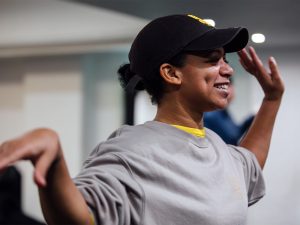 Aria, a black woman with dimples in sports gear smiles as she teaches vogue dance