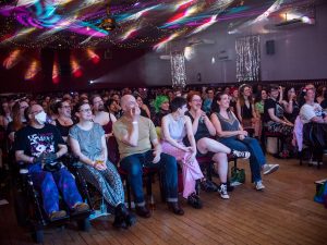 A crowd of people sit looking upwards at a stage in a hall lit by rainbow coloured lights