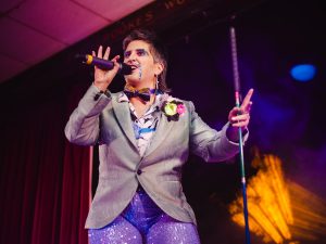 King Tito Bone sings on the mic holding his walking cane. Tito is w hite drag king with a grey mullet and turquoise glitter beard