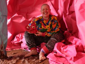 Colour photo, interior. Kitt, a white, shaven headed person with a wide crinkly nosed smile, sits amongst vast sheets of pink and mint green paper crumple to form a cave - like structure around them. on the floor is wet, raw terracotta clay in rolling folds.