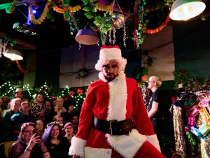 Rhys Pieces a mixed race muscular drag artist with close cut beard is posing to camera dressed in santa costume with the audience behind them